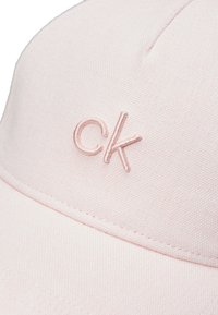 Pursi rožinė kepurė, pagaminta iš tekstūruoto audinio, su išsiuvinėtu „ck“ logotipu priekyje. Iškraipytas šydas užbaigia dizainą.