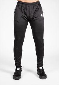 Pantalon de sport noir avec une coupe fuselée, dotés de panneaux latéraux blancs, de poches zippées et d'une texture lisse et extensible.