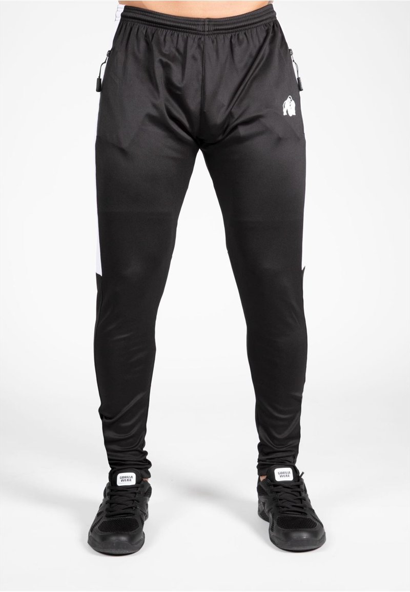 Pantalon de sport noir avec une coupe fuselée, dotés de panneaux latéraux blancs, de poches zippées et d'une texture lisse et extensible.