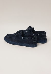 Navy blue suede shoes with a flat sole, lace-up design, and metal eyelets. Features a woven texture around the sole and logo detail.