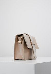 Beige kožená ramenní taška s klopou, nastavitelným popruhem se zlatou sponou, vystavená na bílé podložce na jednobarevném pozadí.