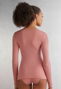 Langsleeve, aansluitend top in zachtroze stof. Glad textuur met een ronde halslijn en een rechte zoom. Achteraan is er een minimaal ontwerp te zien.