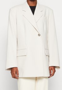 Bílý oversized blejzr s ostrými klopami, jedním zapínáním na knoflík, dvěma předními kapsami a jemnými šicími detaily. Látka působí hladce a elegantně.