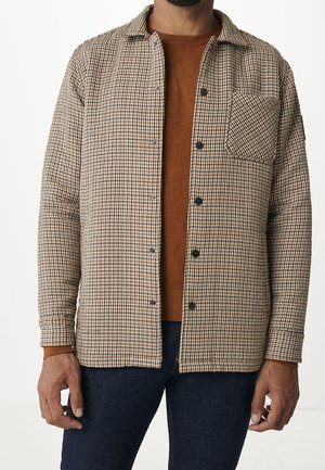 Homme portant une chemise à carreaux marron boutonnée par-dessus un pull couleur rouille avec un pantalon bleu foncé.