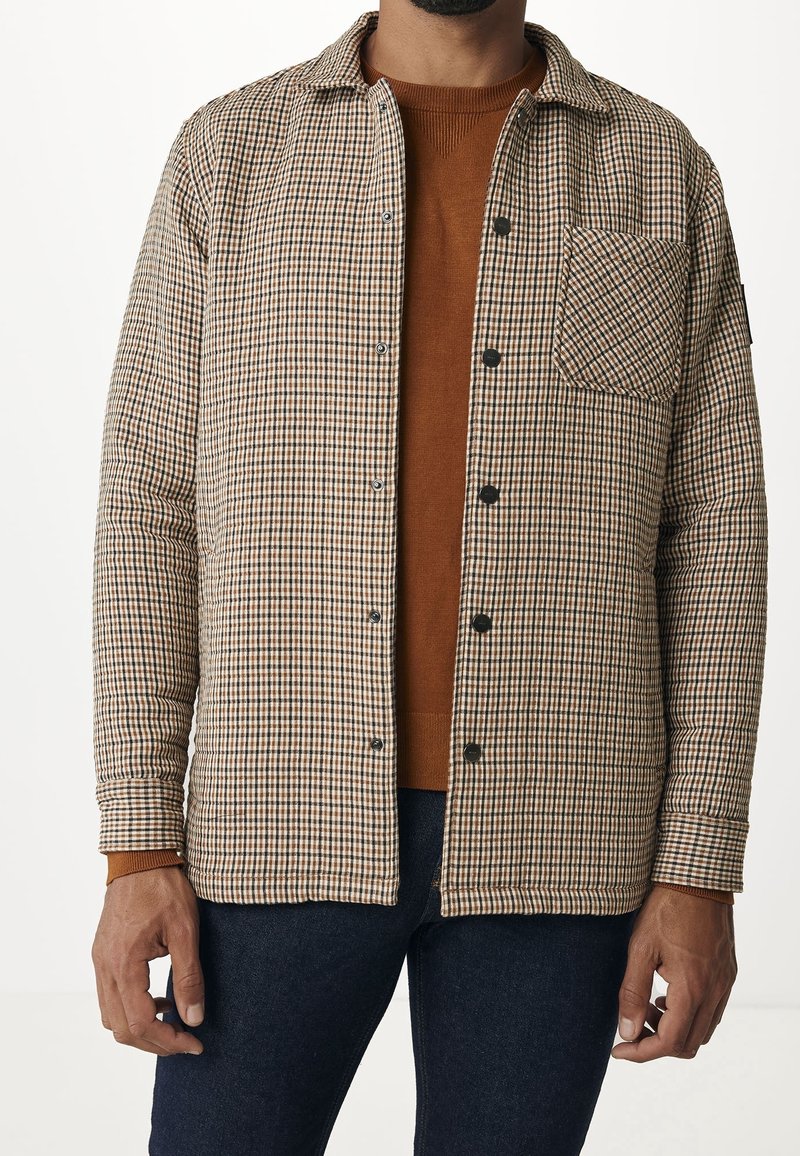 Homme portant une chemise à carreaux marron boutonnée par-dessus un pull couleur rouille avec un pantalon bleu foncé.