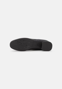 Semelle de chaussure noire avec surface texturée, présentant un bout et un talon lisses, une semelle à motif et un logo central proéminent.
