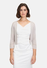 Rövid, világos bézs kardigán háromnegyedes ujjakkal és nyitott elülső résszel. Finom textúrájú részletekkel rendelkezik az ujjak végén.