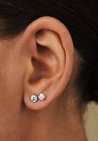 Silberne Ohrstecker mit zwei runden, klaren Edelsteinen unterschiedlicher Größe, vor einem menschlichen Ohr im Close-up.