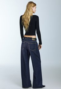 Traperice širokih nogavica u tamnoplavoj boji s glatkom teksturom, s istaknutim stražnjim džepovima i crnim kožnim remenom sa zlatnim detaljem.