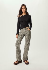Frau steht mit einem schwarzen Off-Shoulder-Oberteil mit Metallösen, Leopardenmuster-Hosen, großen Creolen und Plateausandalen.