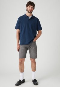 Tummansinisestä puuvillasta valmistettu lyhythihainen polo-paita, yhdistettynä harmaisiin farkkushortseihin. Mustat loafers-kengät ja valkoiset nilkkasukat täydentävät asukokonaisuuden.
