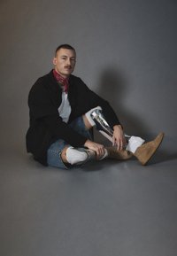 Metalinis protezavimas koja, džinsiniai šortai, balti kojinai, rusvos spalvos kulkšnies batai, juodas per didelis švarkas, raudona skara, ambiente apšvietimas, pilkasis fonas.