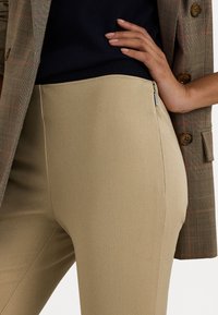 Knee-length beige trousers with a smooth texture, featuring a side zipper, paired with a brown plaid blazer showing button detail.