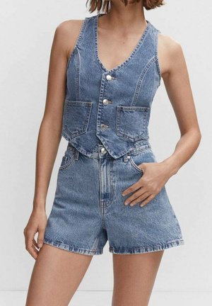 Femme portant un gilet en jean bleu sans manches avec des boutons et un short en jean taille haute assorti, debout avec une main dans la poche.