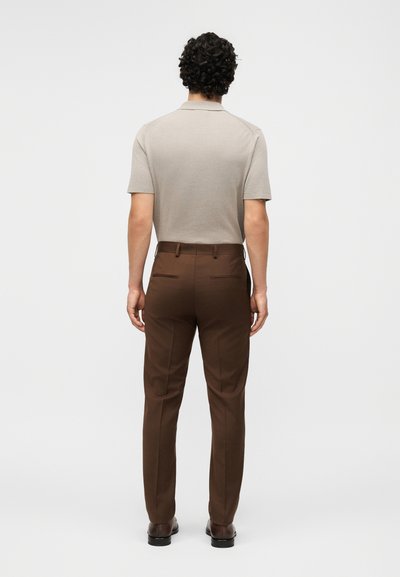 Άνδρας με κατσαρά μαλλιά στέκεται με την πλάτη, φοράει μπεζ κοντομάνικο πουκάμισο, καφέ παντελόνια και σκούρα καφέ παπούτσια.