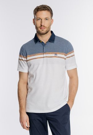 Mężczyzna o krótkich brązowych włosach i brodzie, ubrany w białą, niebieską i pomarańczową koszulkę polo w paski oraz granatowe spodnie, stojący z jedną ręką w kieszeni.