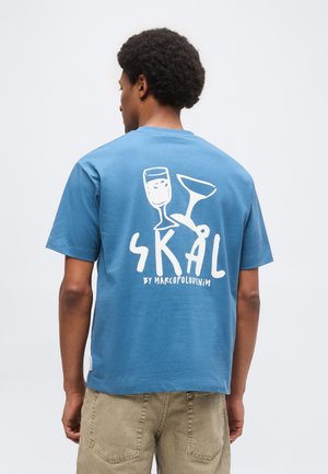 Hombre con camiseta azul con gráfico blanco de dos copas chocando y texto "SKÅL by Marcopolodenim" en la espalda.