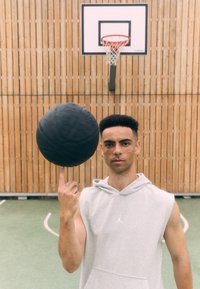 Sort basketball balanceret på en finger, båret af en person i en grå ærmeløs hoodie med et logo. Baggrunden er i træ, og basketballkurven er synlig.