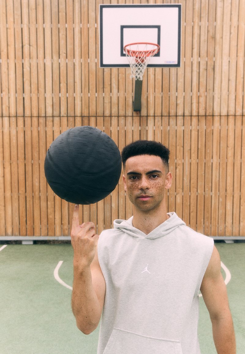 Sort basketball balanceret på en finger, båret af en person i en grå ærmeløs hoodie med et logo. Baggrunden er i træ, og basketballkurven er synlig.