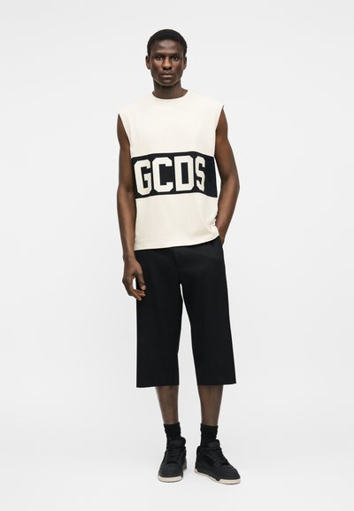 Άνδρας με αμάνικο μπεζ μπλουζάκι με το λογότυπο "GCDS", μαύρο φαρδύ κοντό παντελόνι, μαύρες κάλτσες και μαύρα αθλητικά παπούτσια, στέκεται μπροστά σε λευκό φόντο.