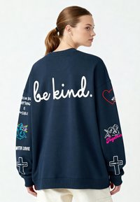 Tumši zila, pārāk liela izmēra sudraba krekls ar uzrakstu "būt laipnam." lielos baltos burtos uz muguras. Piedurknes rotātas ar izdrukātām dizainiem un tekstu, apakšdaļa ar ribota maliņu.