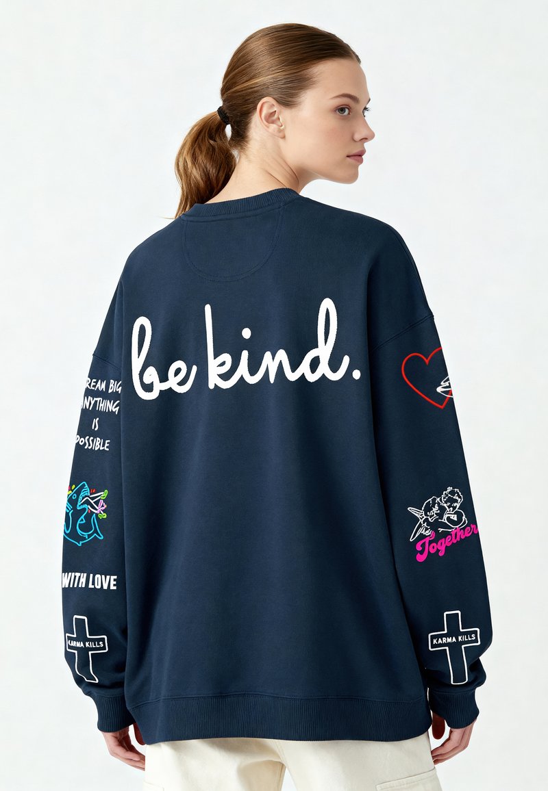 Tumši zila, pārāk liela izmēra sudraba krekls ar uzrakstu "būt laipnam." lielos baltos burtos uz muguras. Piedurknes rotātas ar izdrukātām dizainiem un tekstu, apakšdaļa ar ribota maliņu.