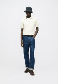 Μπεζ πλεκτό πόλο πουκάμισο με τσέπη στο στήθος, μπλε τζιν τζιν με γυρισμένα μανίκια, καφέ δερμάτινα παπούτσια με κορδόνια και μαύρο καπέλο τύπου κάδο