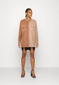 Camisa de camurça em tons de castanho, com fecho frontal em botões, dois bolsos no peito, mangas longas e detalhes em costura branca.