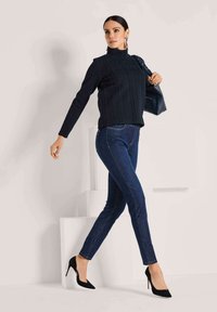 Marinefarbener strukturierten Strickpullover mit hohem Kragen, kombiniert mit dunkelblauen Skinny-Jeans und schwarzen spitzen High Heels, das Model posiert vor einem hellen Hintergrund.