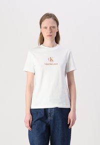 Valkoinen puuvillapaita, jossa on keskeinen oranssi Calvin Klein Jeans -logo, pyöreä kaula-aukko ja lyhyet hihat, yhdistettynä sinisiin farkkuihin.