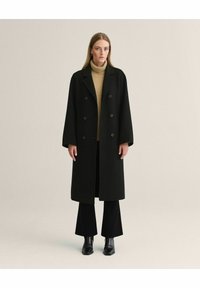 Sort uldfrakke med dobbeltradede knapper, brede revers og en afslappet pasform. Båret med en beige turtleneck og sorte flared bukser.