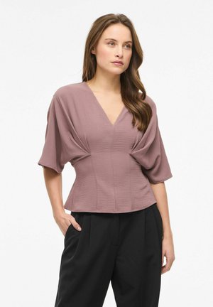 Femme portant un chemisier mauve à col en V avec des manches larges et un pantalon noir taille haute, debout avec une main dans la poche contre un fond blanc.