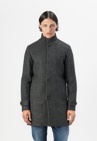 Manteau en laine gris avec un col haut, des boutons sur le devant et des poches latérales. Présente une texture subtile et une coupe ajustée. Porté sur un jean bleu.