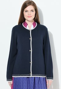 Cárdigan azul marino con cuello redondo, ribete crema y botones plateados. Presenta un dobladillo bordado decorativo. Lleva puesto sobre una camisa con cuello de lunares.