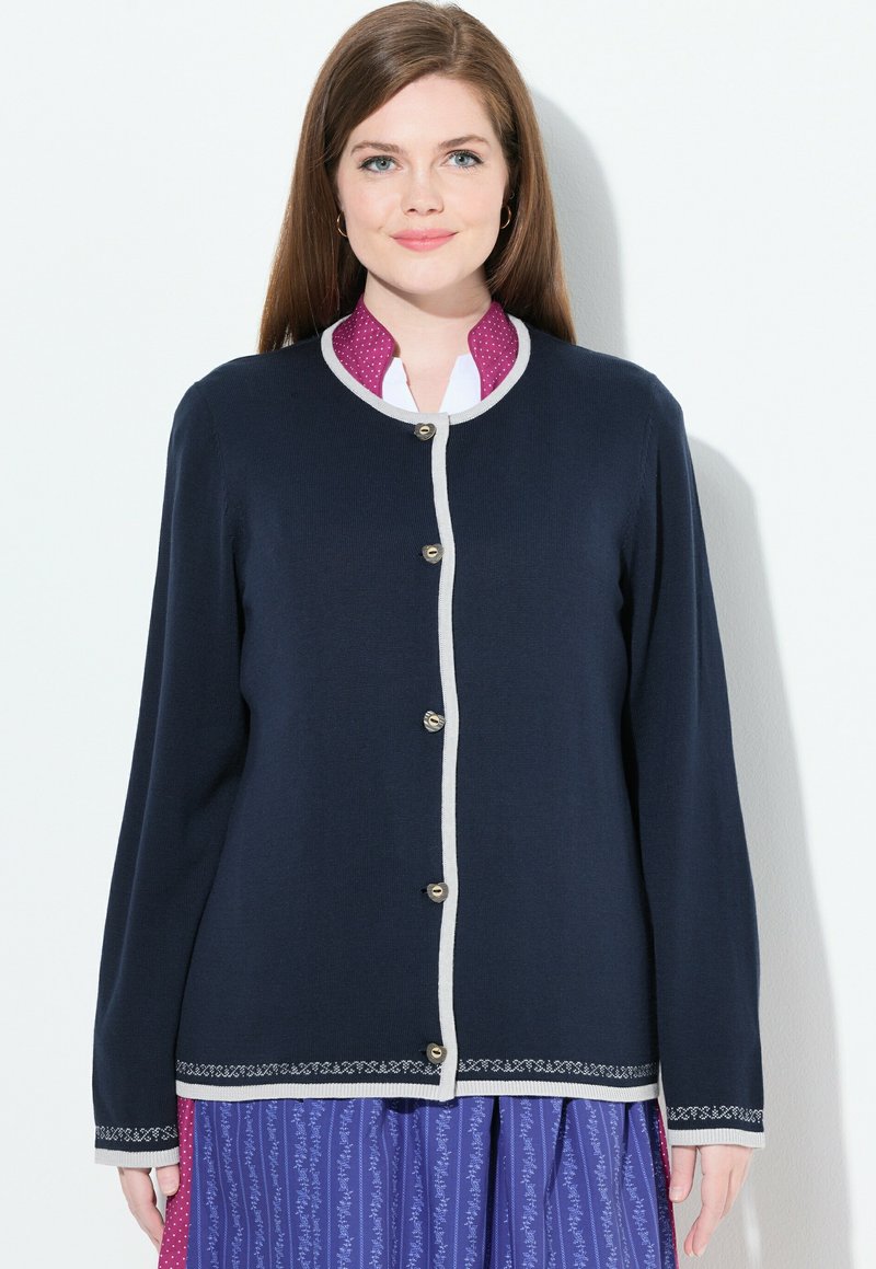 Cárdigan azul marino con cuello redondo, ribete crema y botones plateados. Presenta un dobladillo bordado decorativo. Lleva puesto sobre una camisa con cuello de lunares.