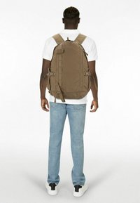 Sac à dos en nylon marron avec des bretelles ajustables, des poches latérales et une texture lisse ; modèle portant un jean bleu clair et une chemise blanche.