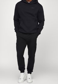 Haut à capuche noir avec logo sur la poitrine, associé à un pantalon cargo noir avec poches latérales et baskets blanches. Tissu lisse avec poignets côtelés.