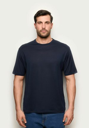 Uomo con capelli corti scuri e barba, indossa una maglietta blu scuro semplice e jeans blu, in piedi contro uno sfondo chiaro.