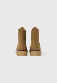 Botas de tobillo de ante marrón con una textura suave, puntera redondeada y suela de goma baja. Cuentan con paneles laterales elásticos y lengüetas para ponerlas y quitarlas fácilmente.