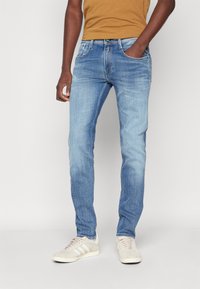 Ljusblå denimjeans med smal passform; har fem fickor, subtil blekning och en klassisk knapp- och dragkedjeöppning, bärs med en brun skjorta.