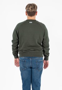 Felpa verde scuro con polsini e orlo a coste, maniche raglan e una piccola etichetta sulla parte superiore della schiena. Abbinata a jeans in denim blu.