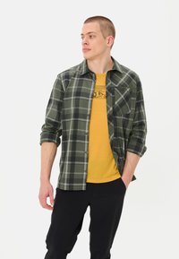 Groene en zwarte geruite flanellen shirt met opgerolde mouwen, gelaagd over een gele graphic t-shirt, gecombineerd met zwarte broek.