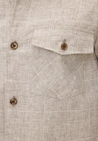 Chemise en tissu à carreaux beige à l'avant avec deux boutons en bois et une poche à rabat boutonnée sur la poitrine.