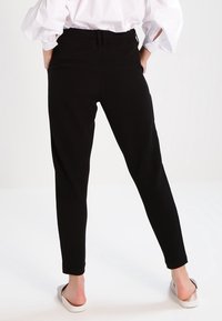 Persoon in zwarte tapered broek, witte blouse met pofmouwen en witte instap schoenen, staand met de handen op de heupen, met de rug naar de kijker gekeerd.