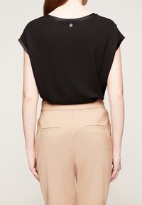 Svart kortärmad topp med rundad halsringning, med en bakknappdetalj, kombinerad med beige byxor och en slät textur.