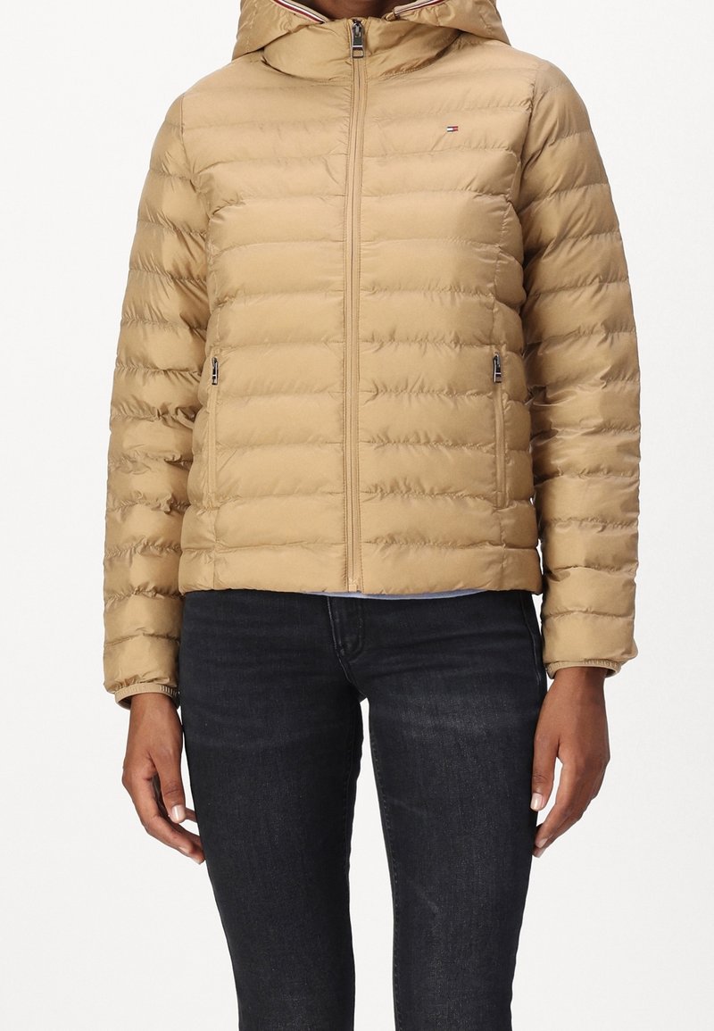 Beżowa kurtka puchowa z zamkiem z przodu, kapturem i poziomym pikowaniem. Posiada boczne kieszenie i detal z logo na piersi.