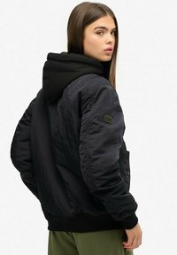 Veste bombardier noire avec capuche, dotée d'une texture lisse, de poignets côtelés et d'une patch sur la manche. Portée sur un pantalon vert olive.