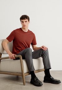 Tricou maron cu guler rotund, pantaloni gri croiți, mocasini negri, șezând pe un scaun din material deschis cu brațe din lemn, pe un fundal simplu.
