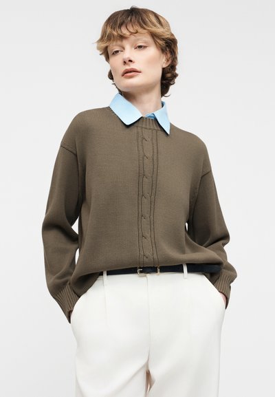 Άτομο που φοράει καφέ πλεκτό ζακέτα με κοτσίδες πάνω από ανοιχτό μπλε πουκάμισο με γιακά, λευκό παντελόνι ψηλής μέσης και μαύρη ζώνη, με τα χέρια στις τσέπες.