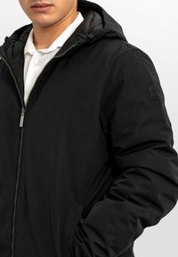 Giacca imbottita nera con chiusura a zip, cappuccio e tasche laterali. Caratterizzata da una texture liscia e un design minimalista. Logo sulla manica.